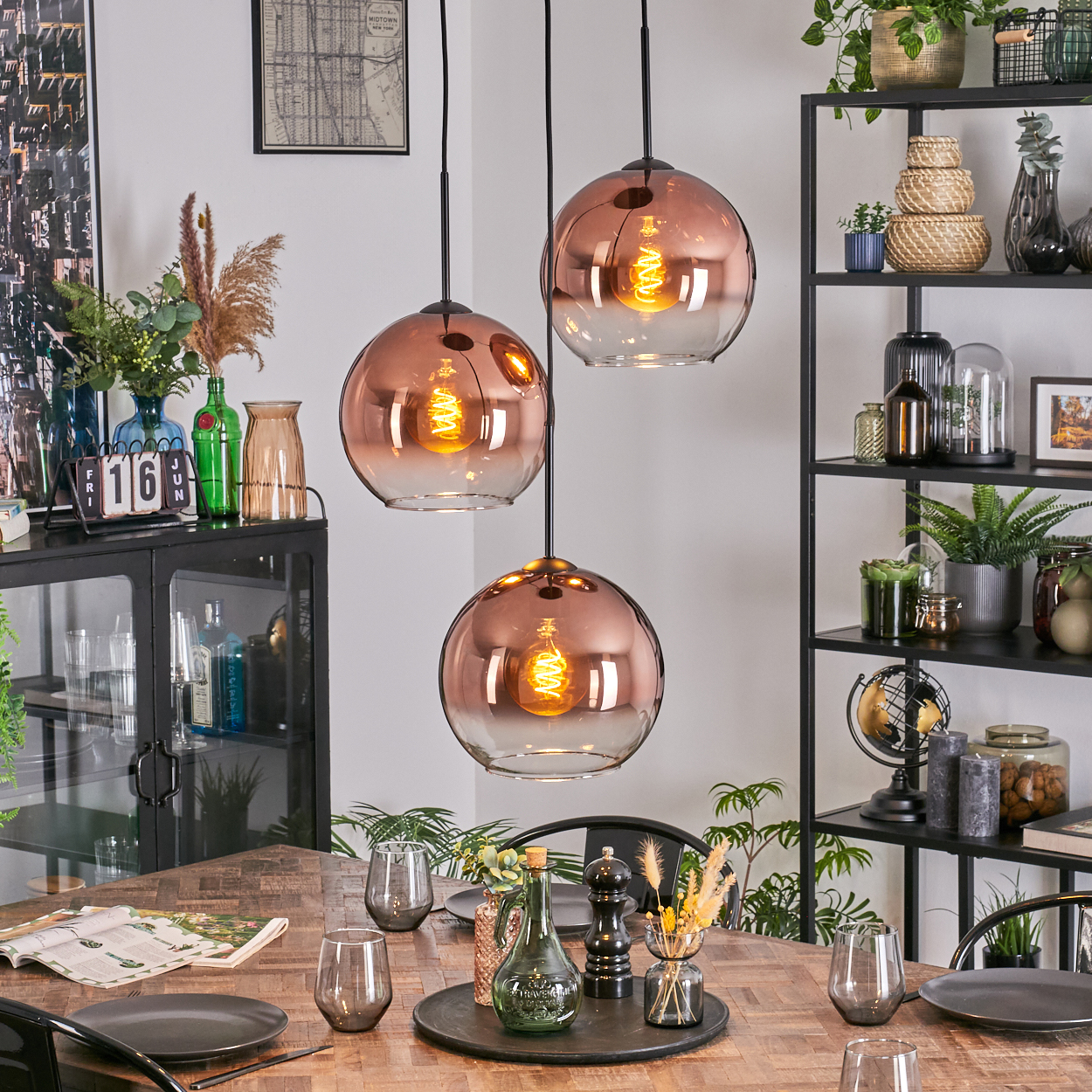 Esszimmer mit Holztisch, Glaslampen, Pflanzen und einem schwarzen Metallregal. Tisch mit Tellern und Gläsern gedeckt.