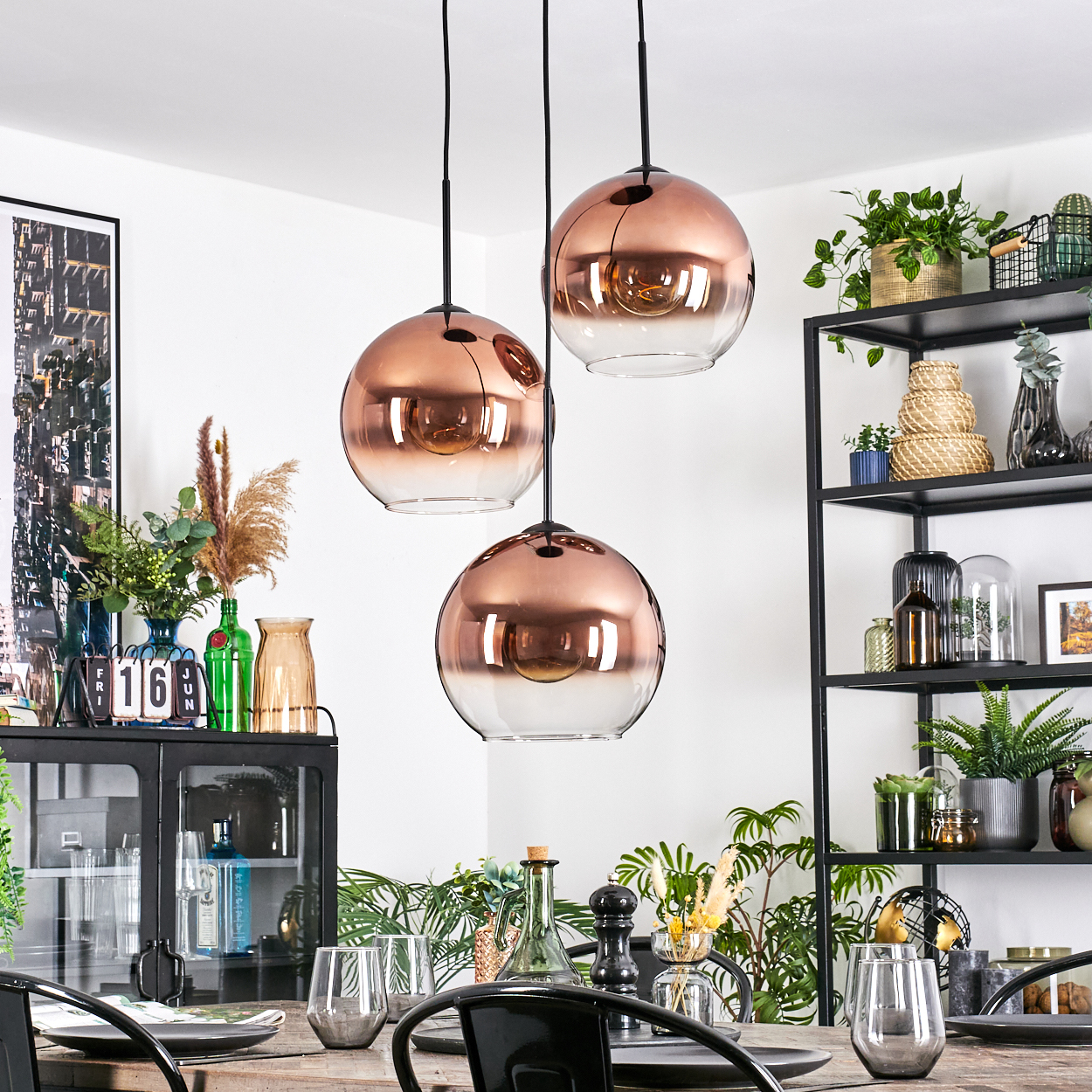 Esszimmer mit Kupfer-Pendelleuchten. Tisch mit Tellern, Gläsern. Regale mit Pflanzen und Dekor. Schwarzer Schrank mit Dekor.