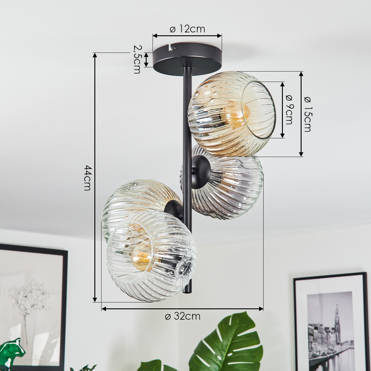 Eine moderne schwarze Pendelleuchte mit vier Glaskugeln, Maße beschriftet. Hintergrund enthält gerahmte Kunst und ein Monstera-Blatt.