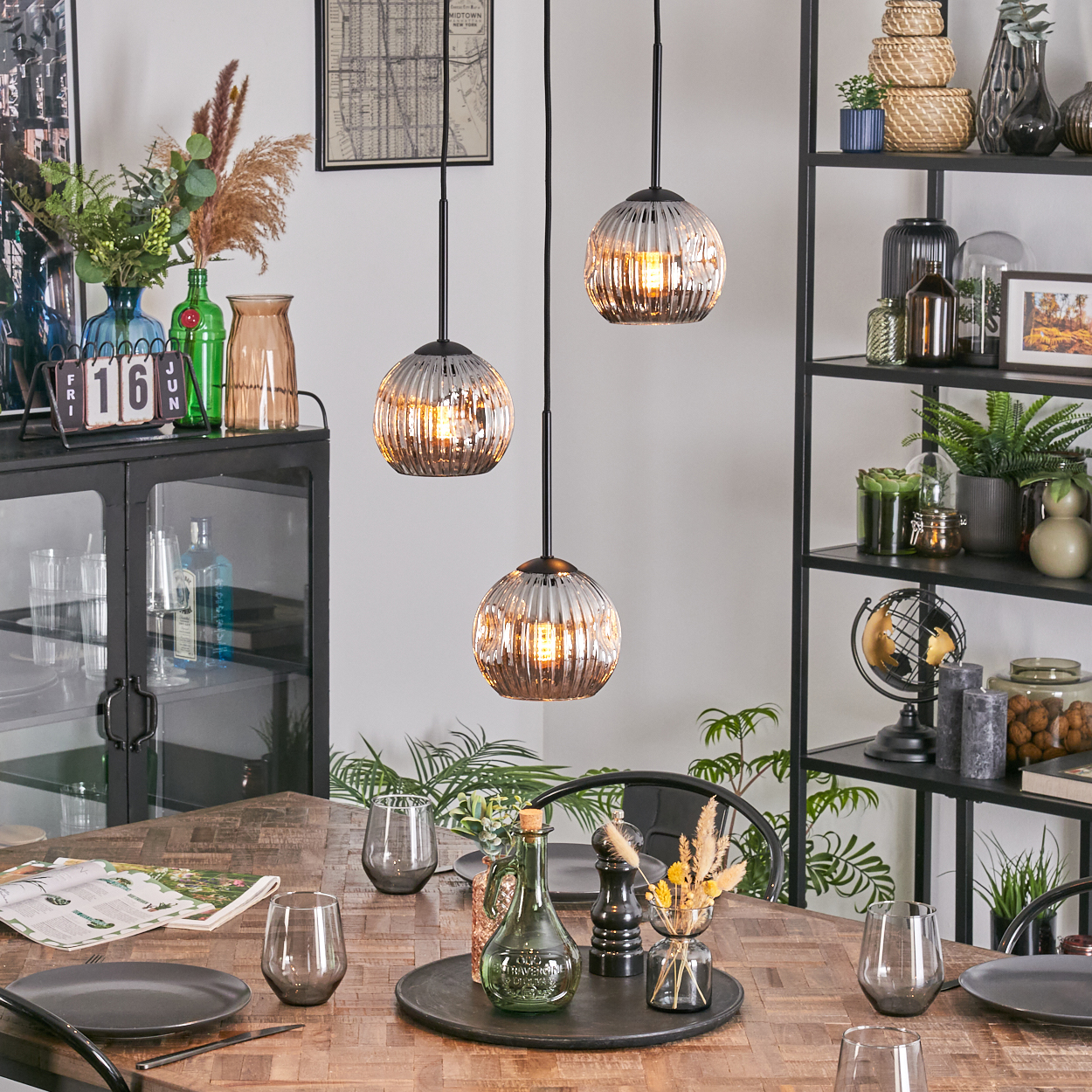 Ein Esszimmer mit einem Holztisch, der zum Abendessen gedeckt ist. Drei Pendelleuchten hängen darüber. Ein schwarzes Regal mit Dekoration im Hintergrund.