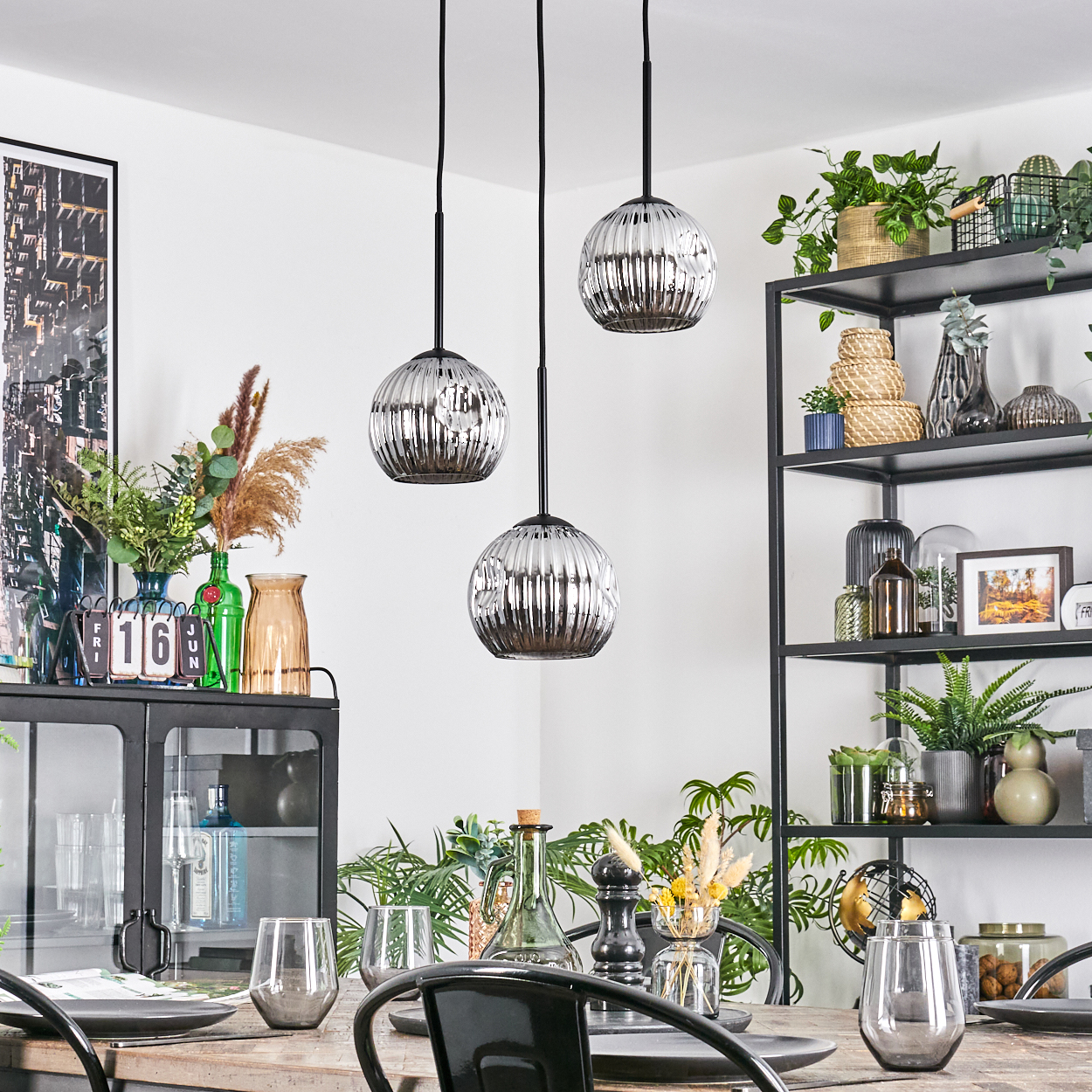 Esszimmer mit drei Hängelampen. Tisch mit Gläsern und Tellern. Regale mit Pflanzen und Dekoration im Hintergrund.