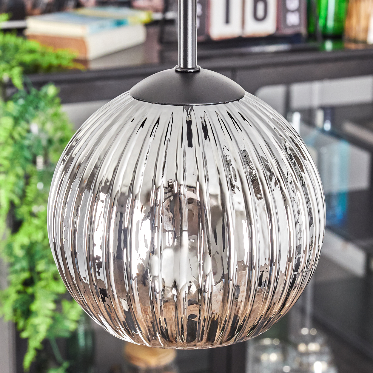 Eine runde, geriffelte Glas-Pendelleuchte hängt. Der Hintergrund zeigt ein Regal, Bücher und grüne Pflanzen. Die Lampe reflektiert das Umgebungslicht.