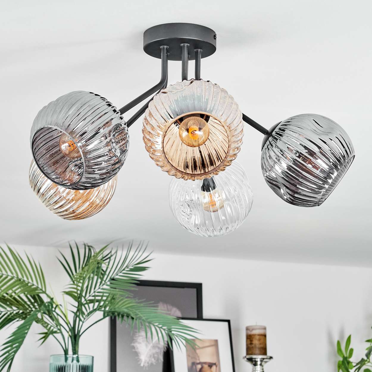 Eine moderne Deckenlampe mit fünf Glaskugeln, grauem Metallrahmen. Darunter eine Pflanze in einer Vase und gerahmte Bilder.