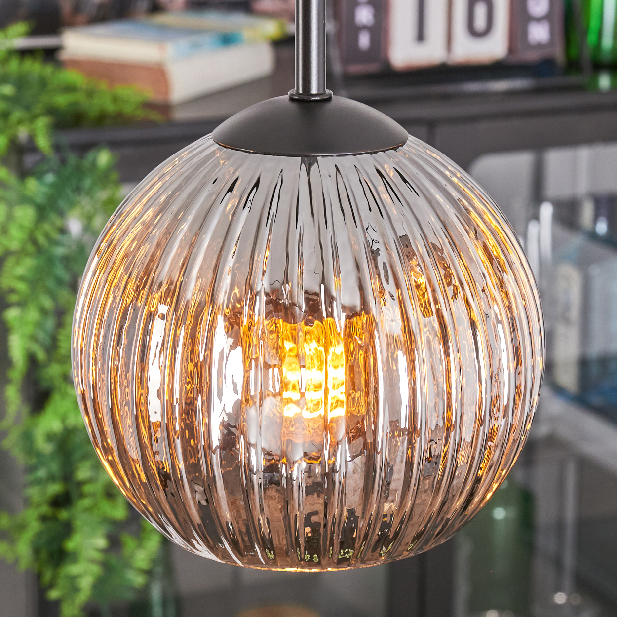 Eine runde, gerippte Glas-Pendelleuchte mit warmer Lichtquelle, die an einer dunklen Halterung hängt. Im Hintergrund grüne Pflanzen und Bücher.