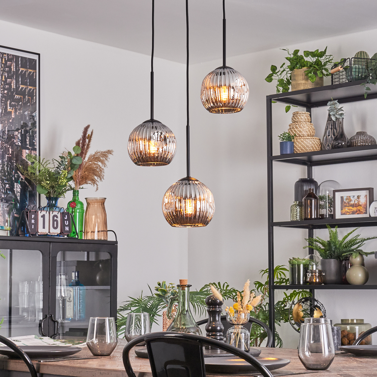 Esszimmer mit drei Hängelampen. Tisch mit Gläsern und Tellern gedeckt. Regale mit Pflanzen und Dekoration im Hintergrund.