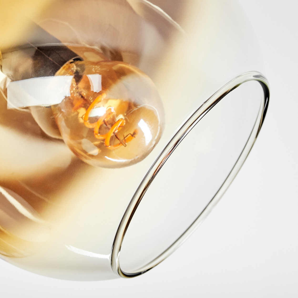 Nahaufnahme eines Glaslampenschirms mit sichtbarer Glühbirne. Die Glühbirne hat einen warmen, orangefarbenen Schein und einen spiralförmigen Glühfaden, vor weißem Hintergrund.