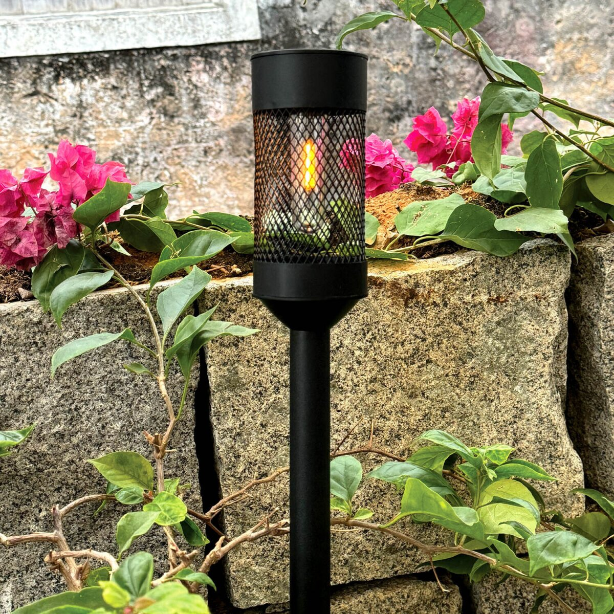 Eine schwarze Fackellampe mit Flammeneffekt steht vor einer Steinmauer. Rosa Blumen und grüne Blätter umgeben die Lampe.