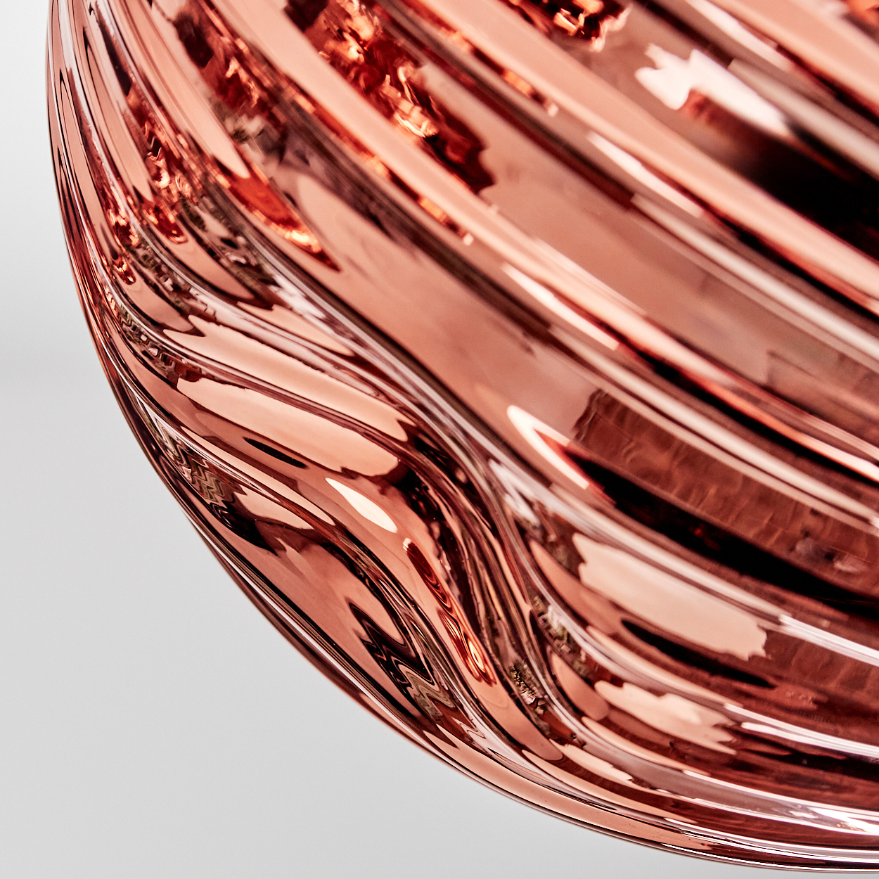 Nahaufnahme eines wellenförmigen, roségoldenen Metallobjekts vor weißem Hintergrund. Reflexionen und Licht spielen auf der gekrümmten Oberfläche.