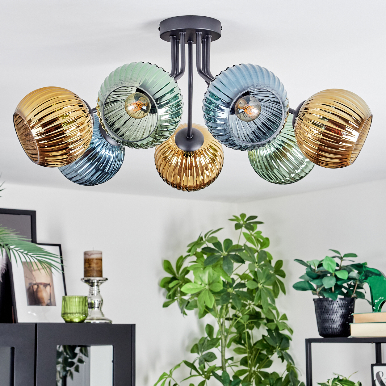 Moderne Deckenlampe mit sechs farbigen Glasschirmen. Der Raum hat Pflanzen, einen Schrank und ein Regal mit Büchern. Die Lampe steht im Mittelpunkt.