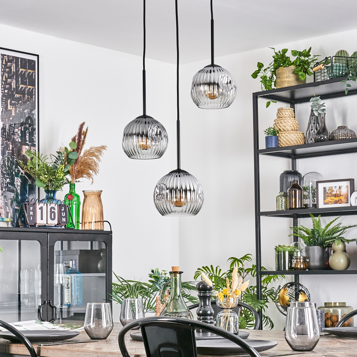 Esszimmer mit drei Hängelampen. Tisch mit Gläsern und Tellern gedeckt. Regale mit Pflanzen und Dekoration.