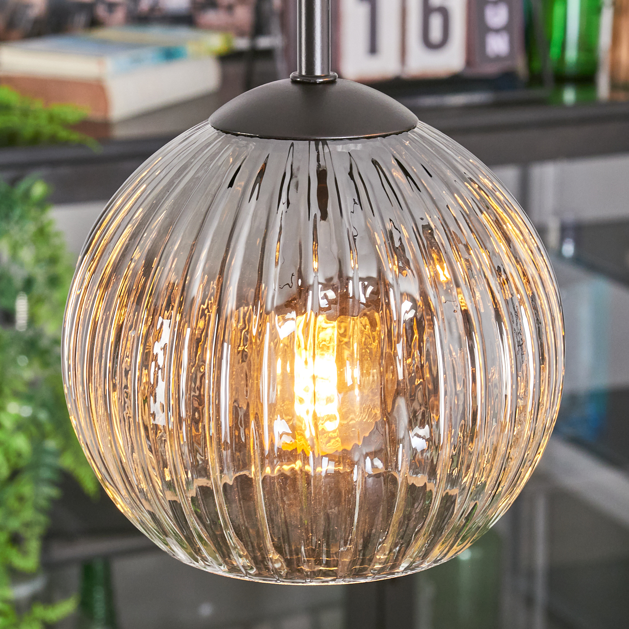 Eine klare, geriffelte Glas-Pendelleuchte mit warmem Leuchtmittel hängt. Hintergrund hat eine unscharfe Ansicht eines Regals und Pflanzen.