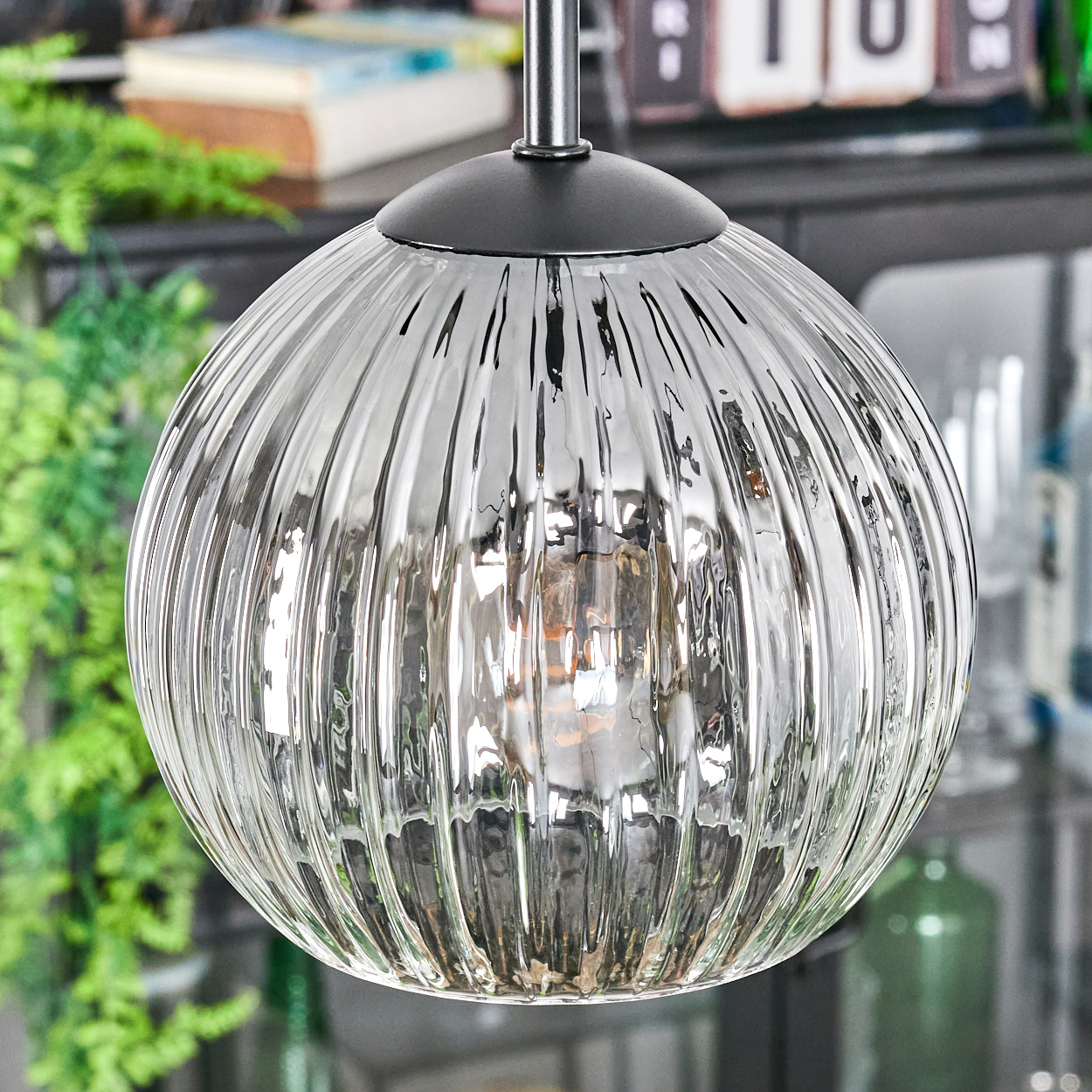 Eine klare, geriffelte Glas-Pendelleuchte hängt von der Decke. Im Hintergrund sind grüne Pflanzen und andere Objekte zu sehen.