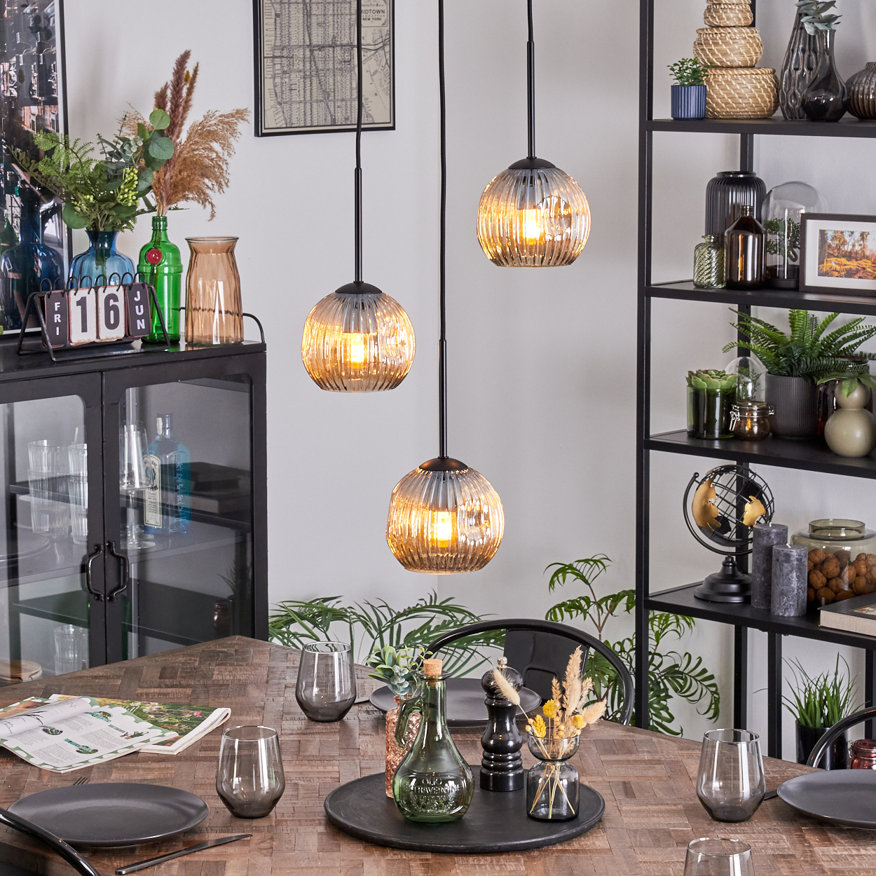 Esszimmer-Szene mit Holztisch, Hängelampen und Dekorationsartikeln auf Regalen. Teller, Gläser und ein Mittelstück stehen auf dem Tisch.