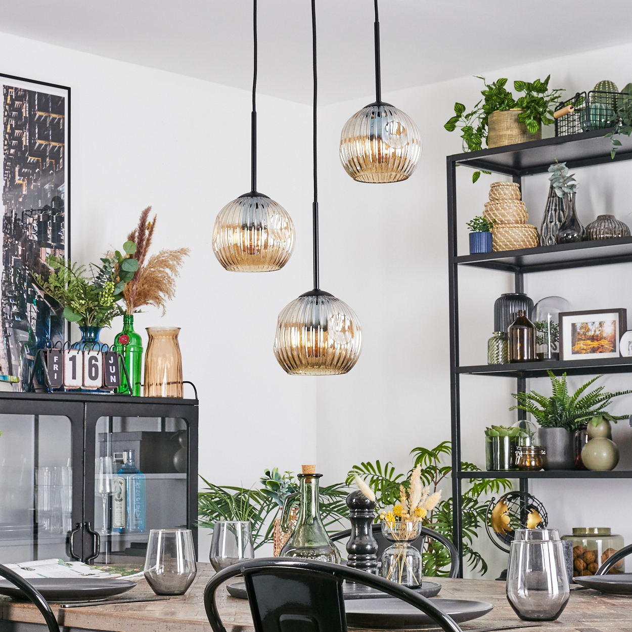 Esszimmer mit drei Hängelampen, Tisch mit Gläsern und Tellern, Pflanzen und einem schwarzen Metallregal mit Dekorationen.