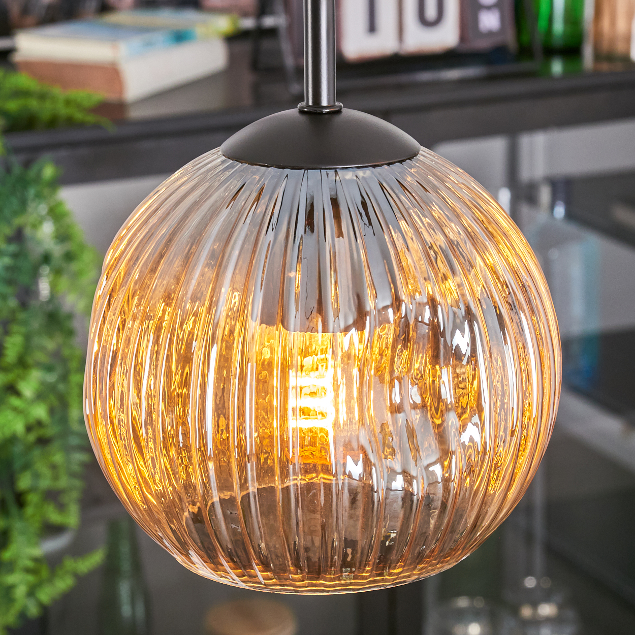 Nahaufnahme einer Pendelleuchte mit geripptem, bernsteinfarbenem Glasschirm. Die Glühbirne leuchtet hell. Der Hintergrund umfasst Grün und Dekoration.