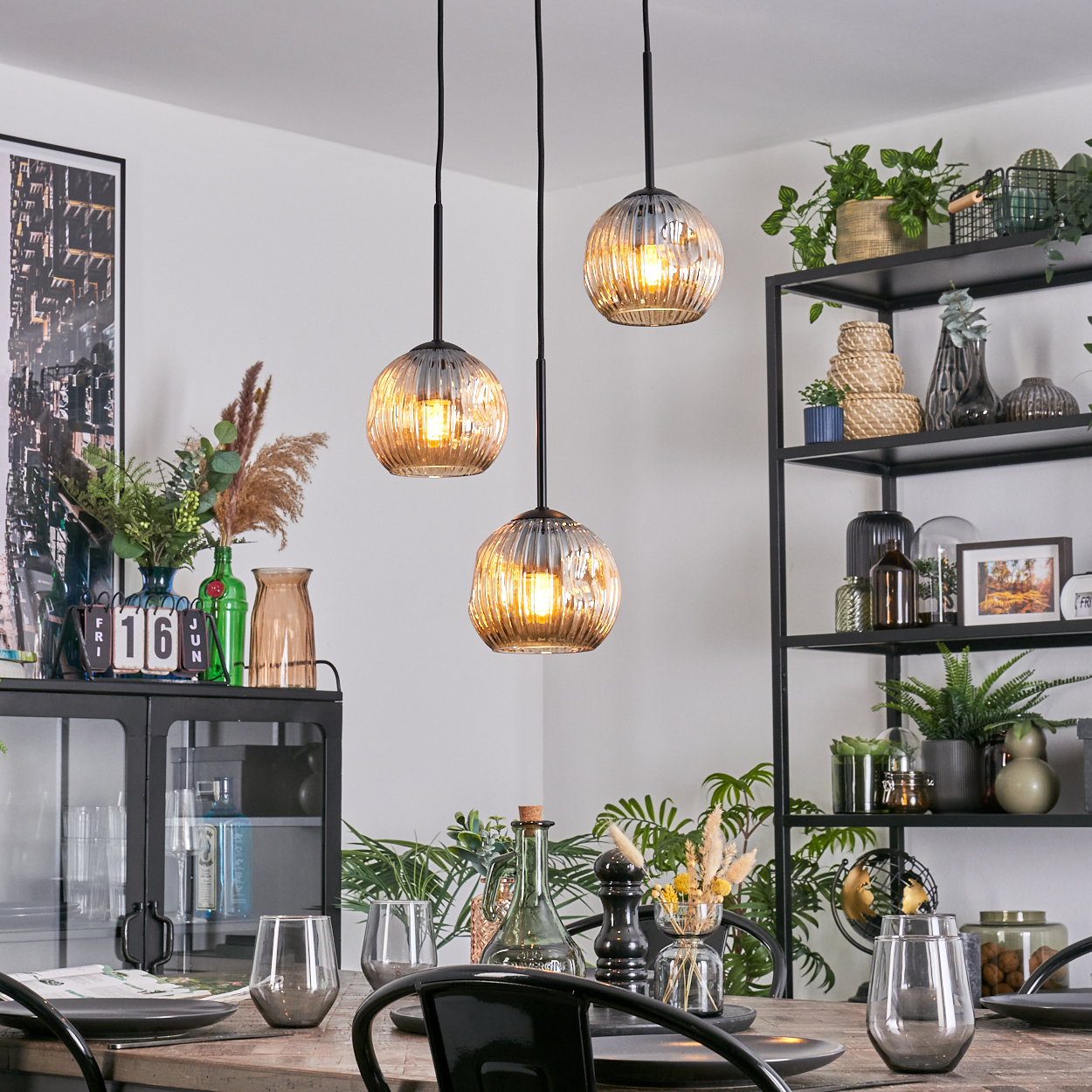 Esszimmer mit drei Pendelleuchten. Tisch mit Gläsern und Tellern gedeckt. Pflanzen und Dekoration auf Regalen und in Vasen.
