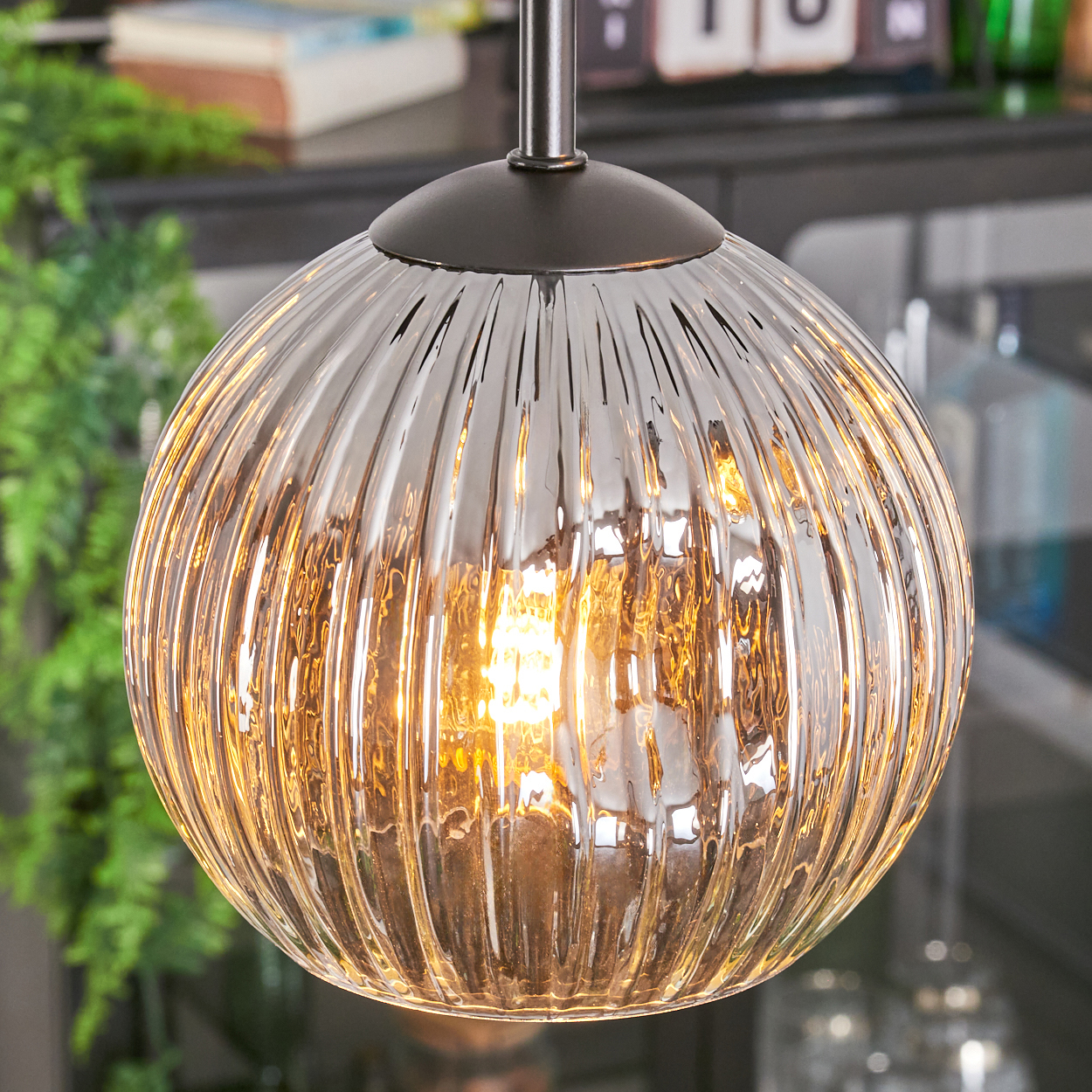 Eine runde Glas-Pendelleuchte mit geripptem Design. Sie hat ein warmes Leuchten von der Glühbirne im Inneren. Im Hintergrund sind grüne Pflanzen.