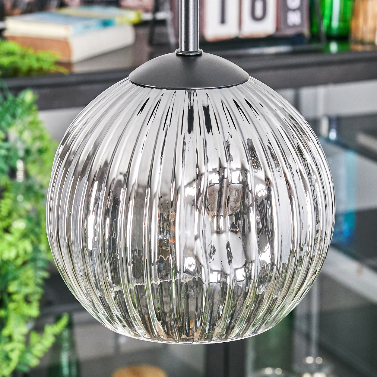 Nahaufnahme einer Glas-Pendelleuchte mit vertikalen Rippen. Grüne Pflanzen und andere Gegenstände sind im Hintergrund unscharf.