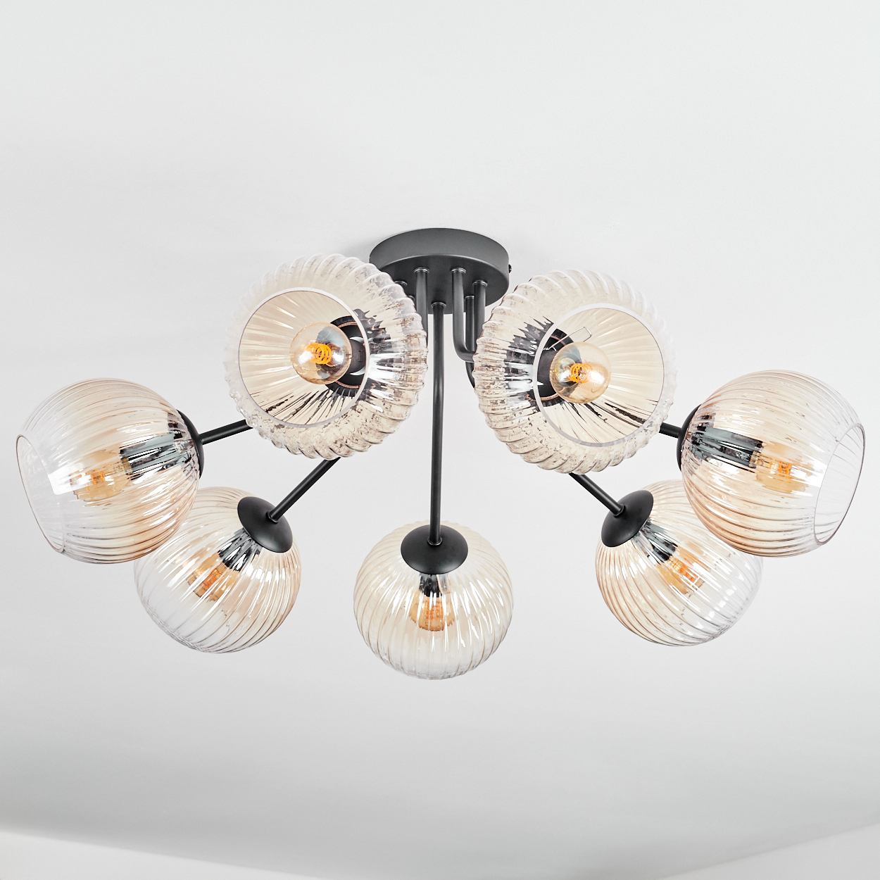 Eine moderne Deckenleuchte mit sechs Leuchten. Jede Leuchte hat einen Glaskolben mit einer warmen Glühbirne und schwarzen Metallarmen.