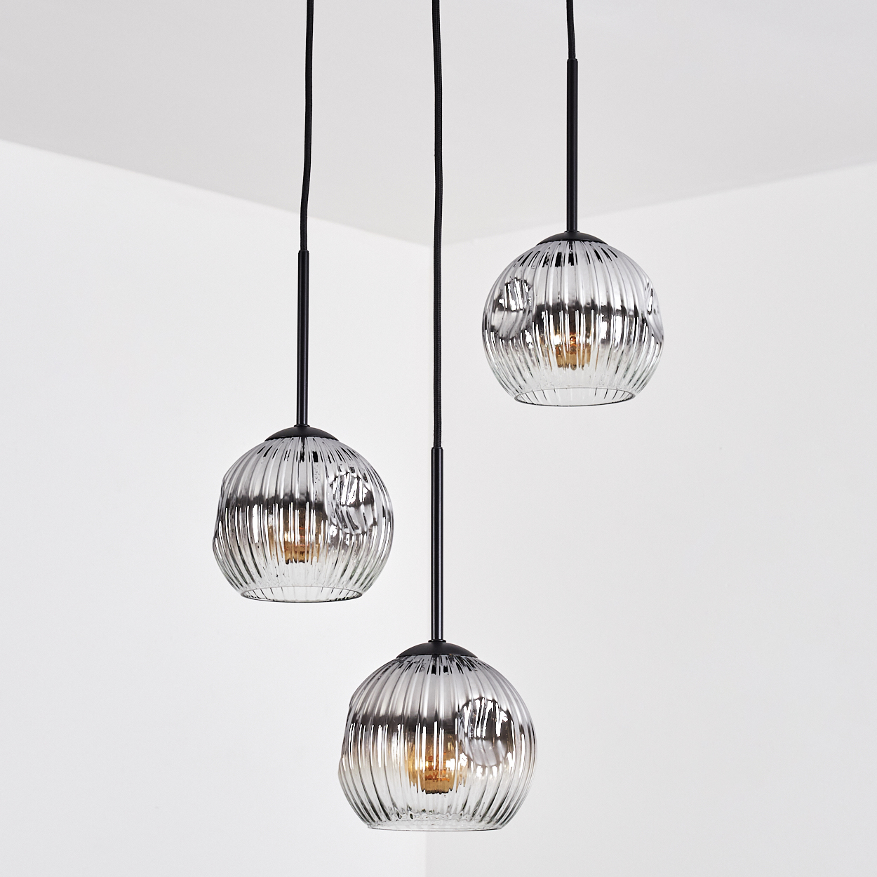 Drei Pendelleuchten mit strukturierten Glasschirmen hängen an einer weißen Ecke. Schwarze Kabel und Armaturen. Die Lichter sind an.