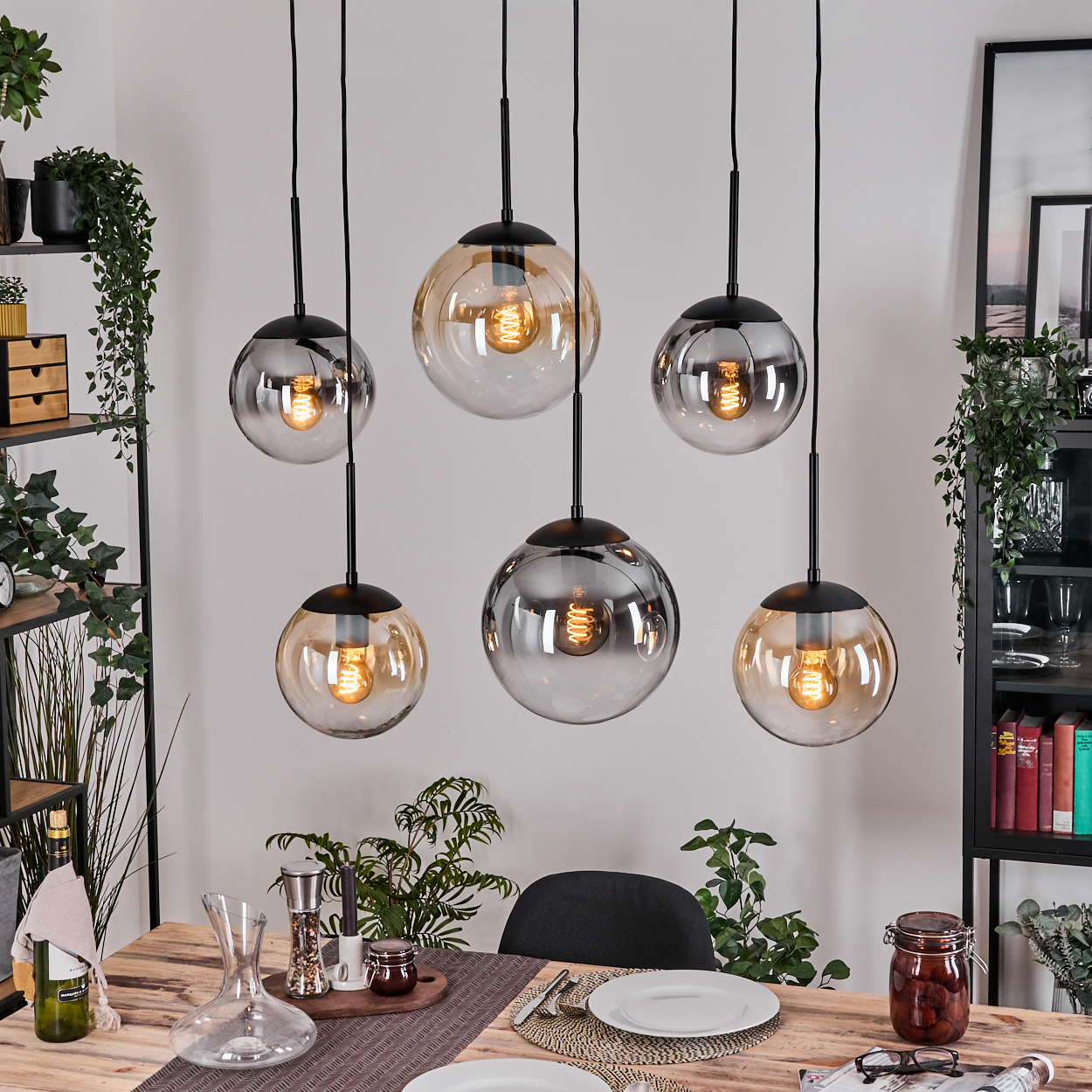 Esszimmer mit sechs hängenden Kugelleuchten. Tisch mit Tellern, Pflanzen und Dekanter gedeckt. Ein Glas, Bücher und Regale sind sichtbar.