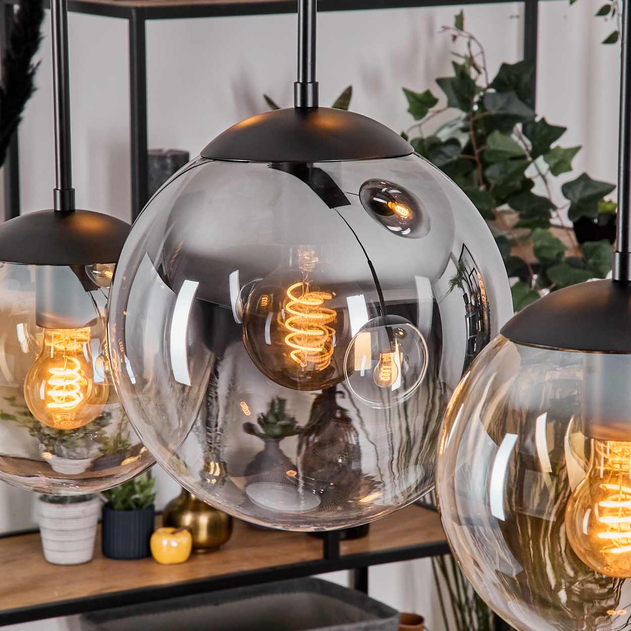 Drei kugelförmige Pendelleuchten mit sichtbaren Glühbirnen. Schwarze Armaturen. Hintergrund zeigt Pflanzen, Regale mit Töpfen und einen goldenen Apfel.