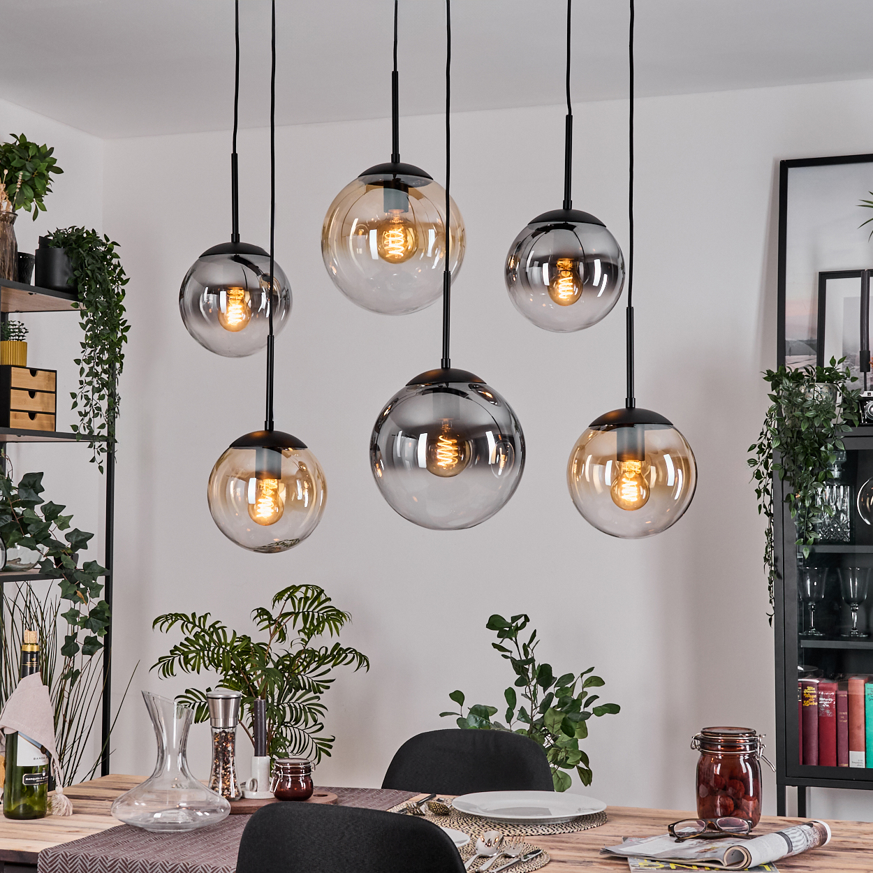 Sechs kugelförmige Pendelleuchten hängen über einem Esstisch. Die Szene umfasst Pflanzen, eine Glaskaraffe und ein Glas mit Essen.