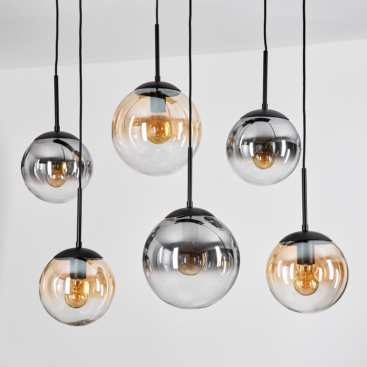 Sechs moderne Pendelleuchten mit Glaskugeln und freiliegenden Glühbirnen hängen vor einer weißen Wand. Einige Kugeln sind verspiegelt.