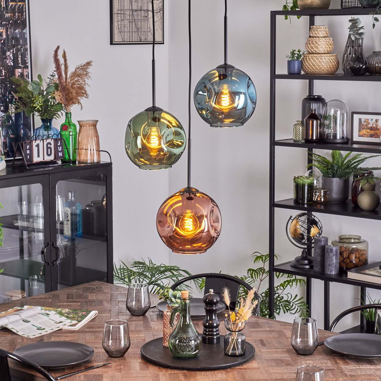 Esszimmer mit Holztisch, drei hängenden Glaslampen und einem schwarzen Regal mit Dekoration. Teller, Gläser und Pflanzen sind vorhanden.