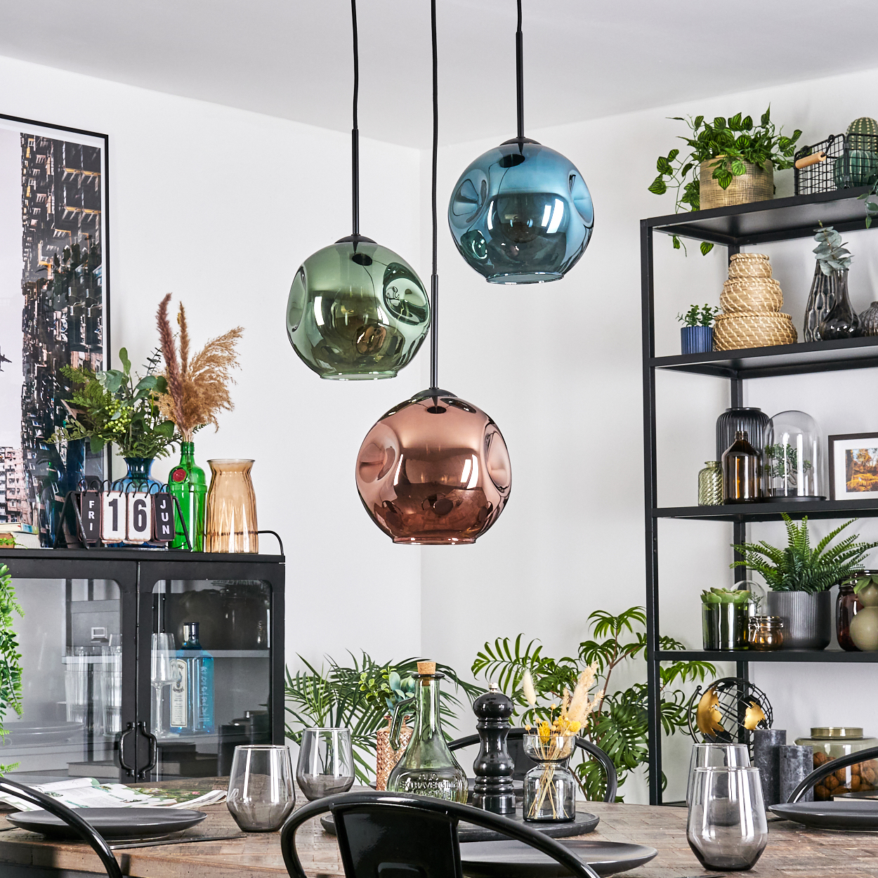 Drei farbige Pendelleuchten hängen über einem Holztisch. Regale mit Pflanzen und Dekor sind im Hintergrund.