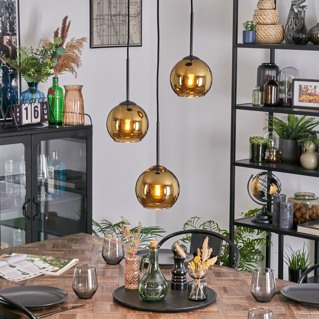 Esszimmer mit Holztisch, goldenen Pendelleuchten, schwarzem Regal mit Deko und einem Kalender an der Seite.