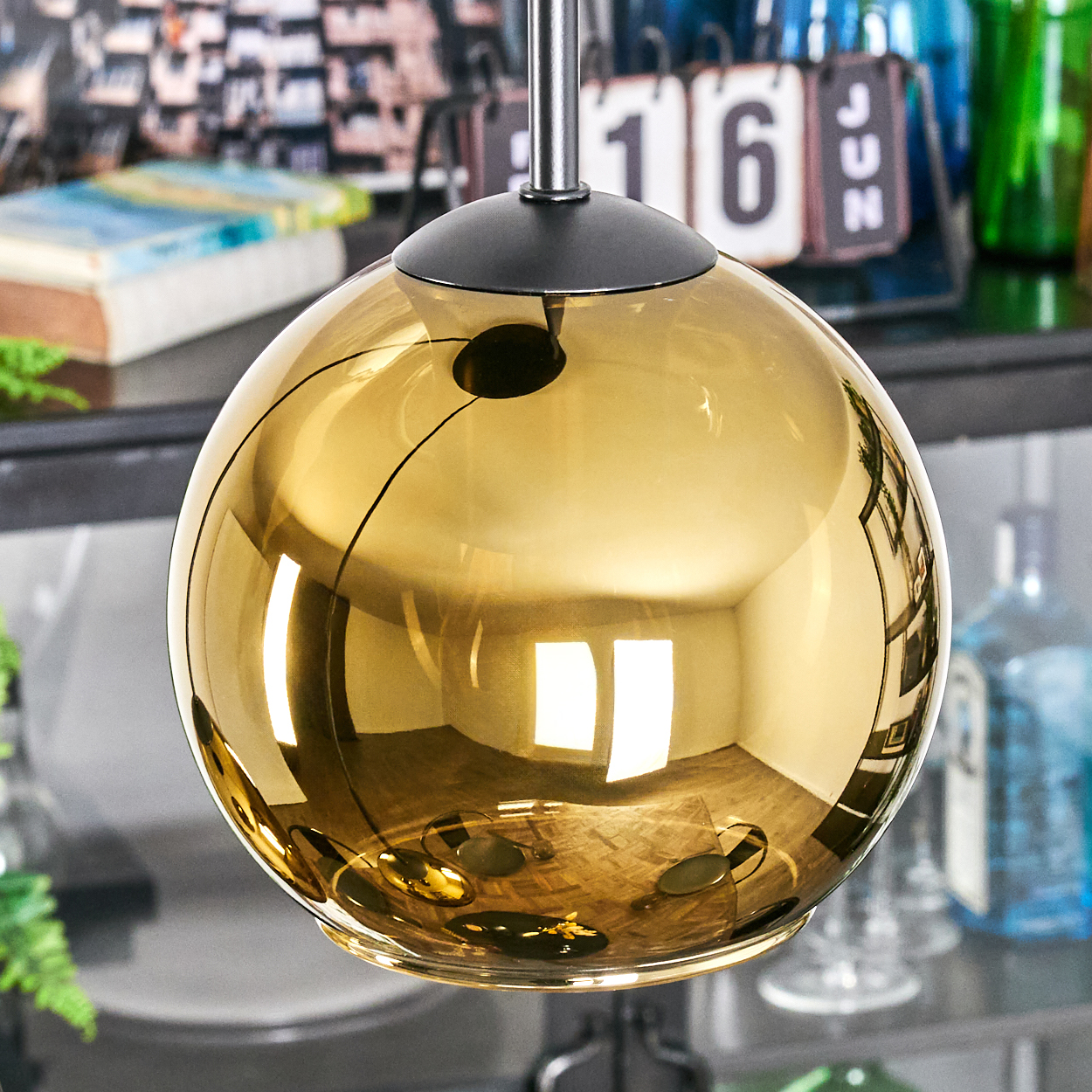 Eine goldene, kugelförmige Lampe hängt. Sie reflektiert den Raum, einen Kalender und andere Objekte. Die Lampe hat einen schwarzen Deckel und eine silberne Stange.