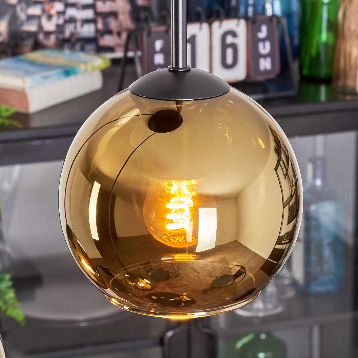 Eine goldene, kugelförmige Pendelleuchte mit einer leuchtenden Glühbirne. Der Hintergrund umfasst einen Kalender, Bücher und grüne Glasflaschen.
