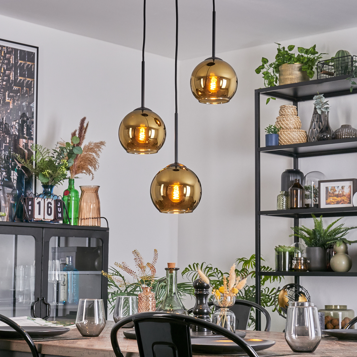Esszimmer mit drei Hängelampen. Tisch mit Gläsern und Tellern gedeckt. Regale mit Pflanzen und Dekor im Hintergrund.