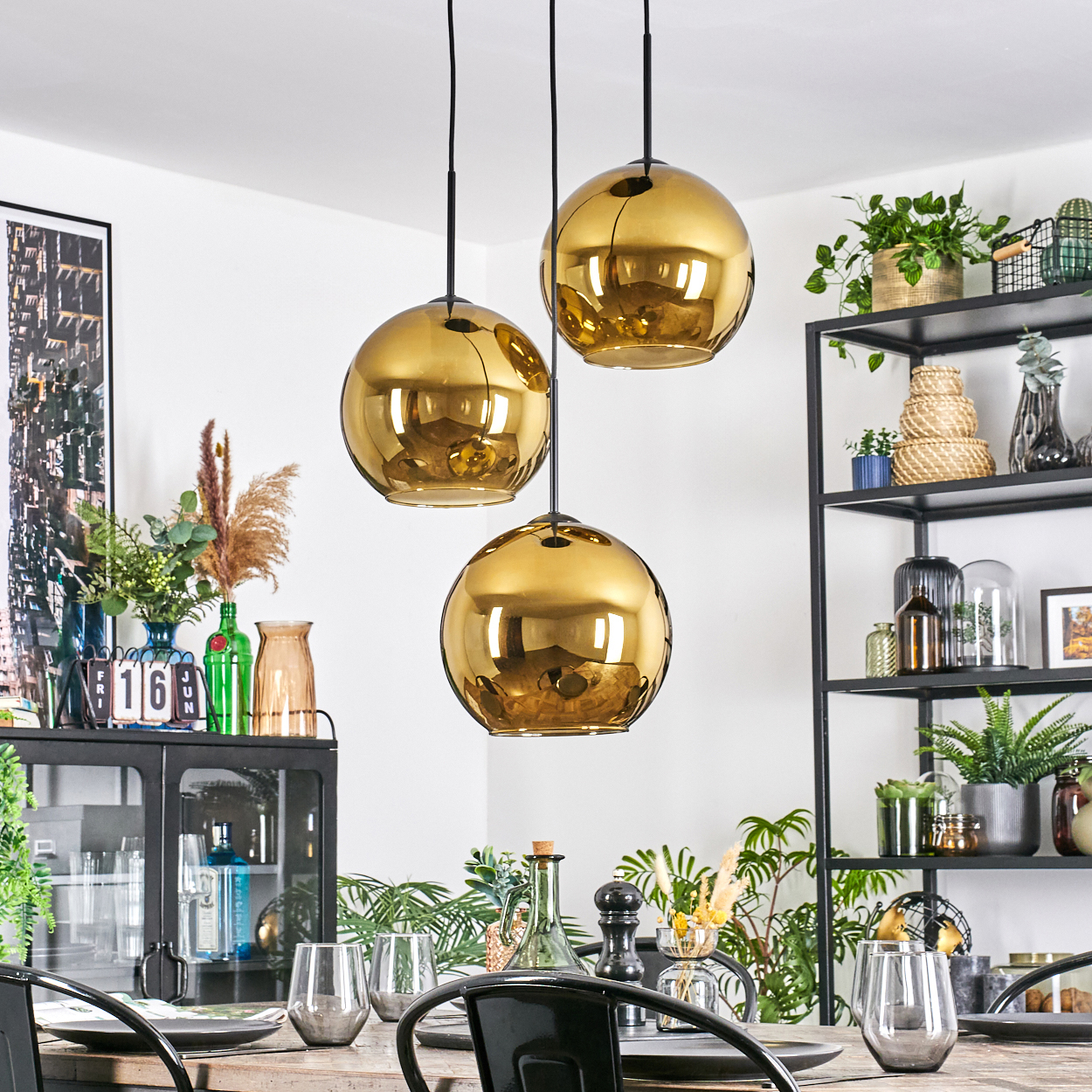 Drei goldene Pendelleuchten hängen über einem Holztisch, der zum Abendessen gedeckt ist. Regale mit Pflanzen, Vasen und ein Schrank im Hintergrund.