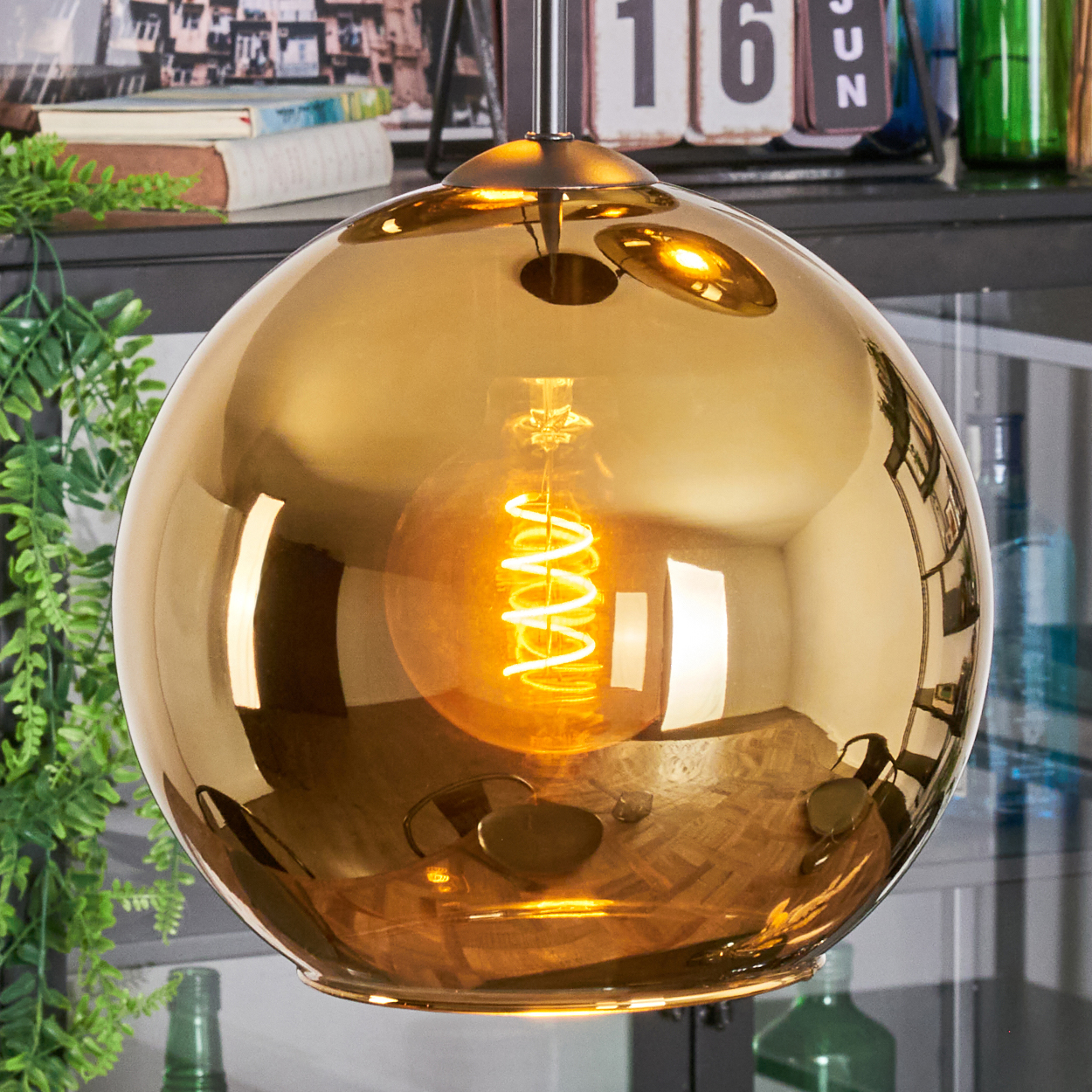 Eine goldene, kugelförmige Hängelampe mit einer leuchtenden, spiralförmigen Glühbirne. Der Hintergrund zeigt Regale und Grün, mit einem Kalender.