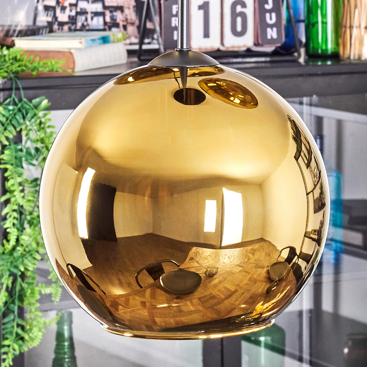 Eine goldene, kugelförmige Lampe spiegelt einen Raum wider. Im Hintergrund sind grüne Pflanzen und ein Kalender zu sehen.