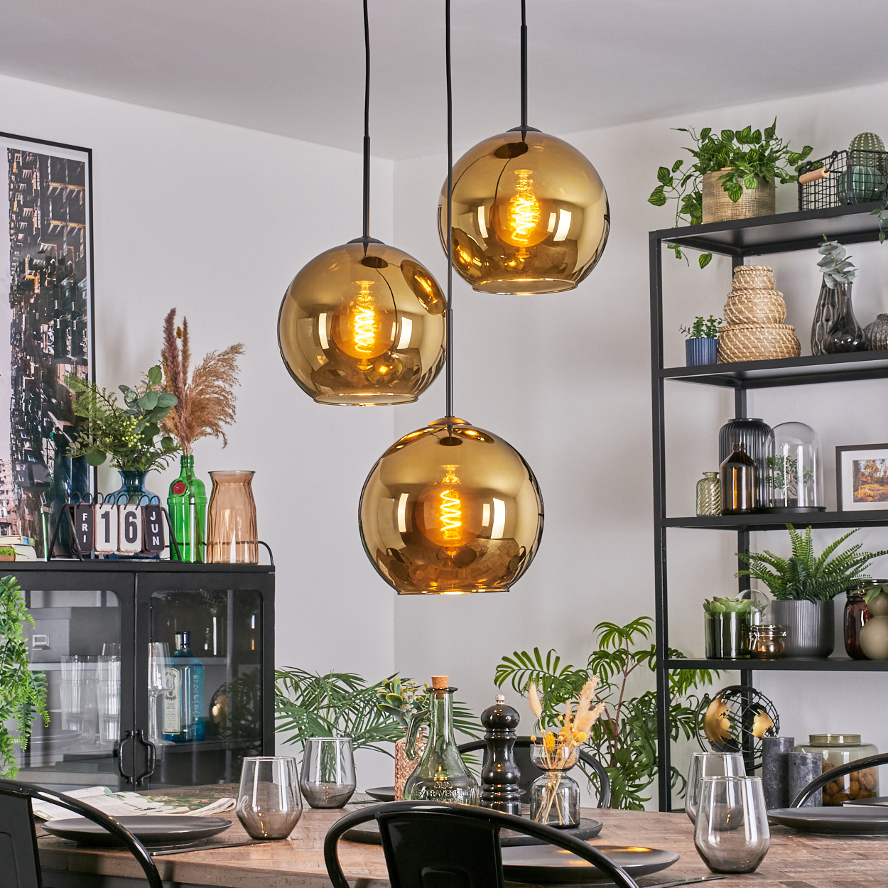 Esszimmer mit drei goldenen Pendelleuchten. Tisch mit Gläsern und Tellern gedeckt. Regale mit Pflanzen und Dekoration.