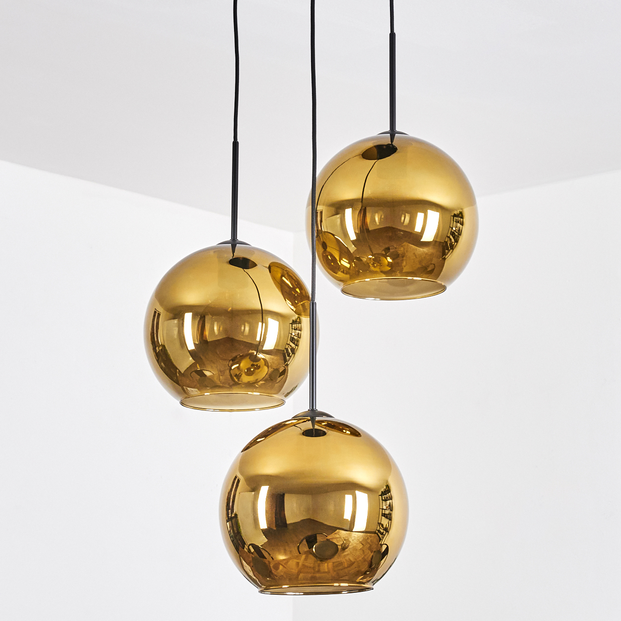 Drei goldene, kugelförmige Pendelleuchten, die an schwarzen Schnüren an einer weißen Wand hängen. Die Lichter reflektieren den Raum.