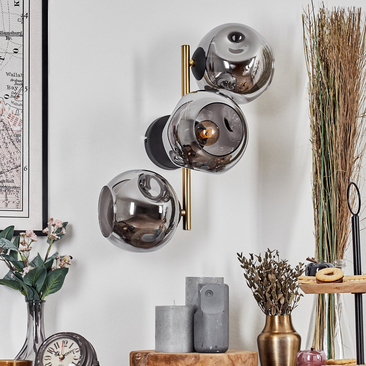 Eine moderne Wandlampe mit drei Glaskugeln. Darunter Kerzen, eine Vase und eine Uhr auf einer Holzoberfläche. Links hängt eine Karte.