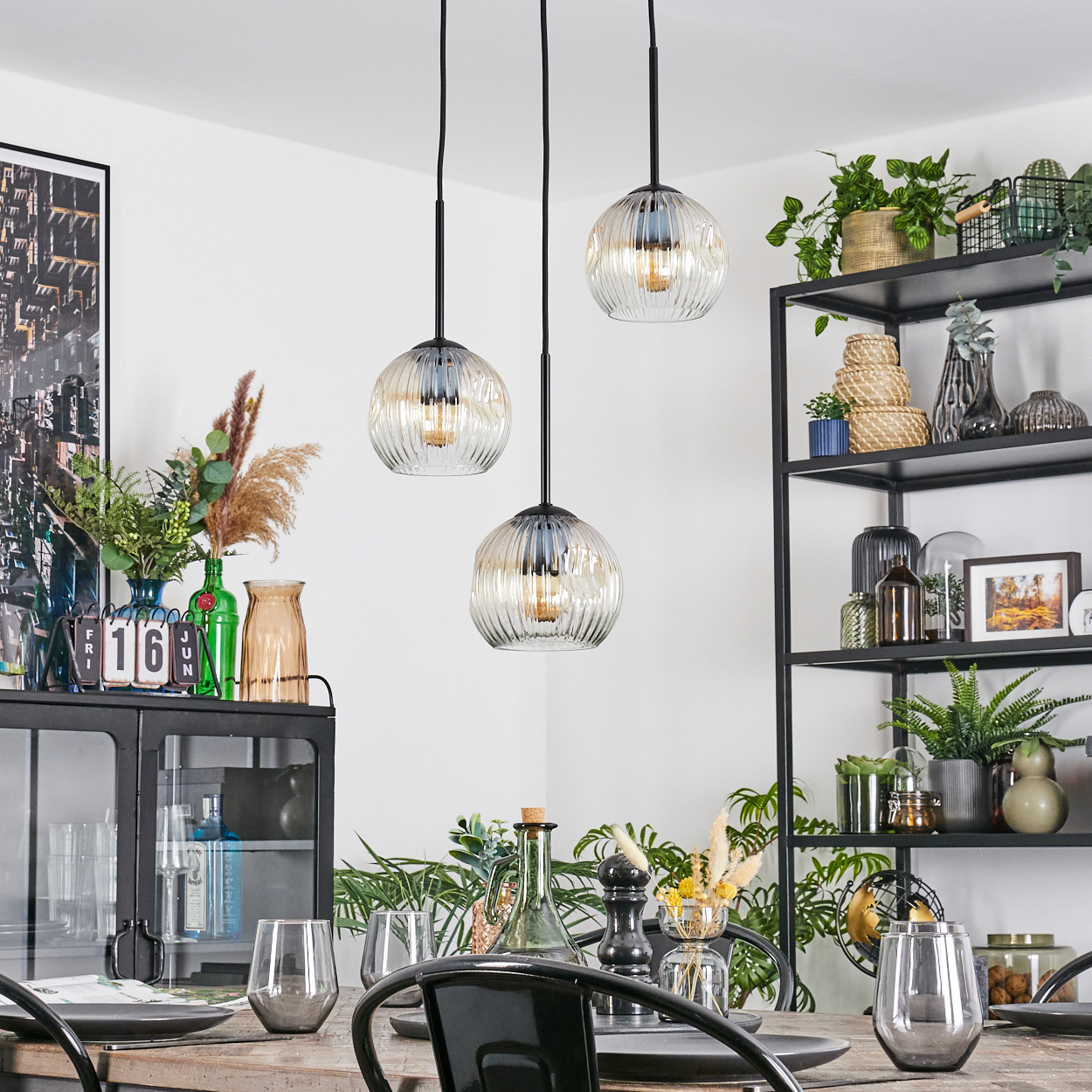 Esszimmer mit drei Pendelleuchten. Tisch mit Gläsern und Tellern gedeckt. Regale mit Pflanzen und Dekoration im Hintergrund.