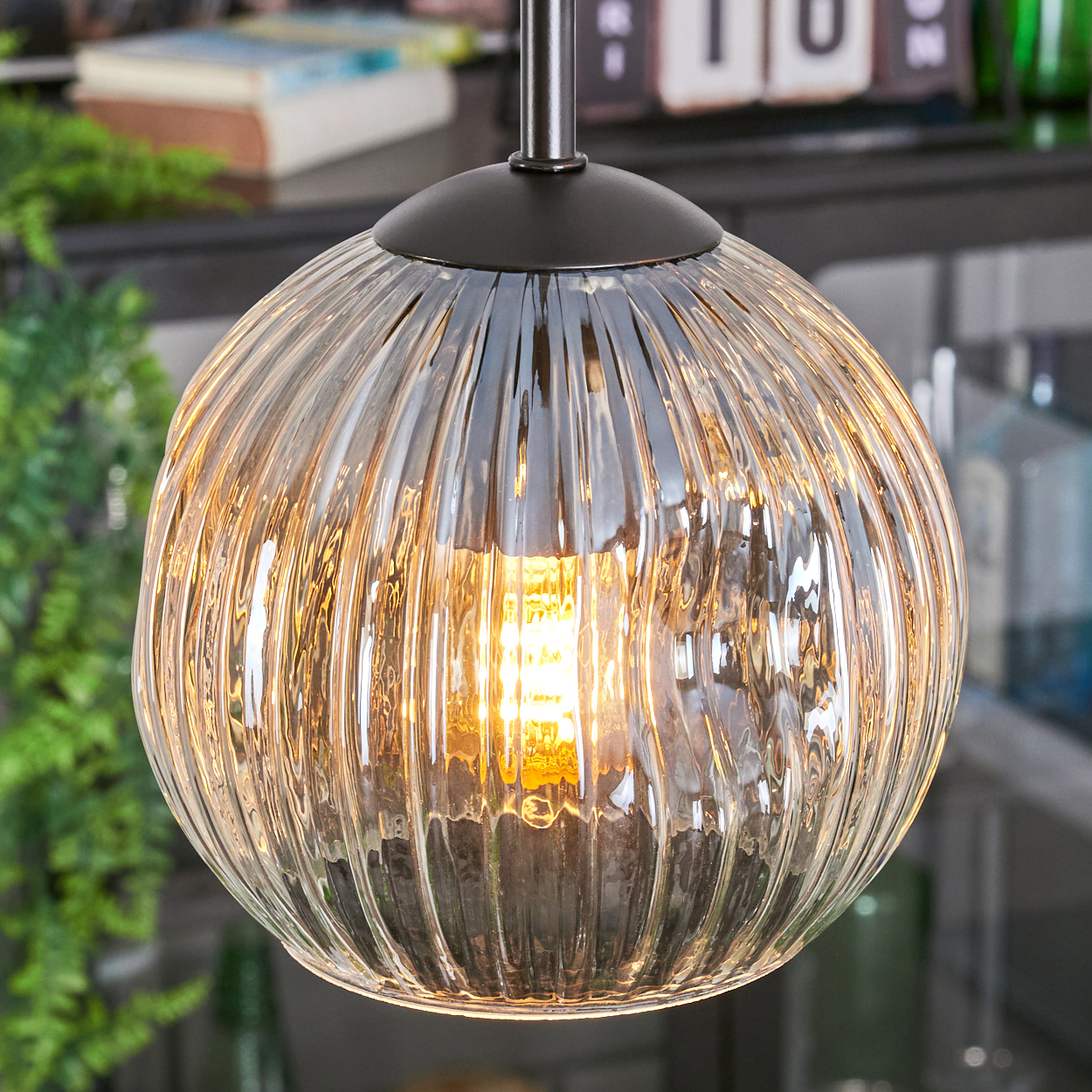 Eine klare, gerippte Glas-Hängeleuchte mit einer leuchtenden Glühbirne. Der Hintergrund zeigt ein verschwommenes Interieur.