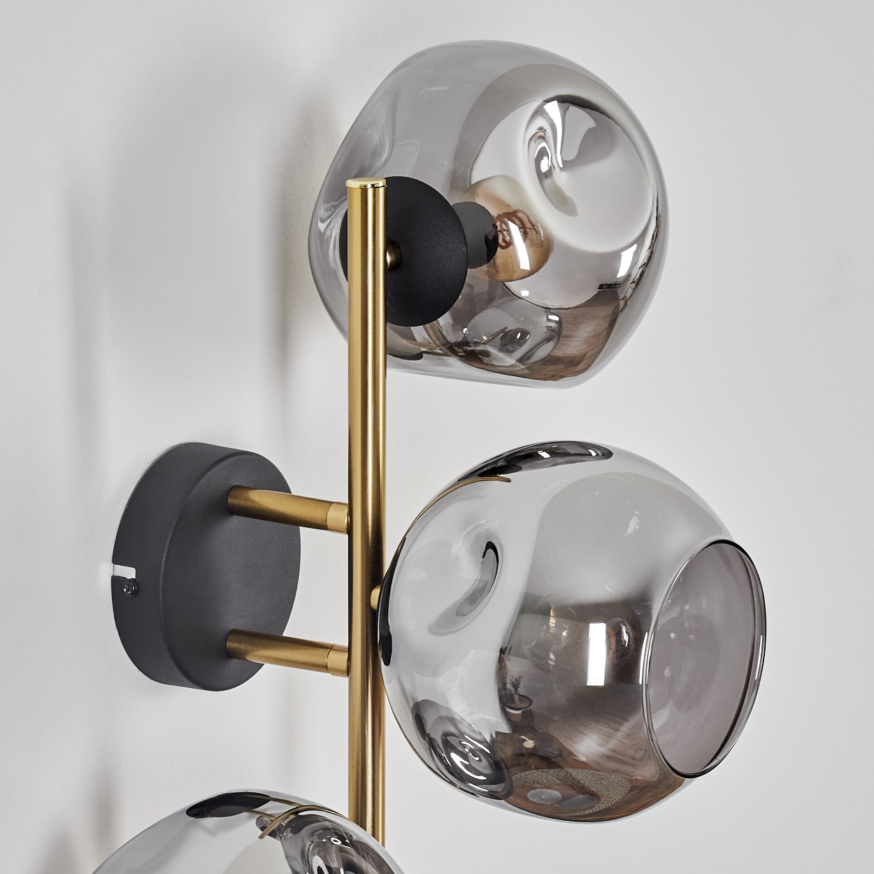 Eine moderne Wandlampe mit drei grauen Glaskugeln. Die Lampe hat einen goldenen Rahmen und einen schwarzen Sockel, montiert an einer weißen Wand.