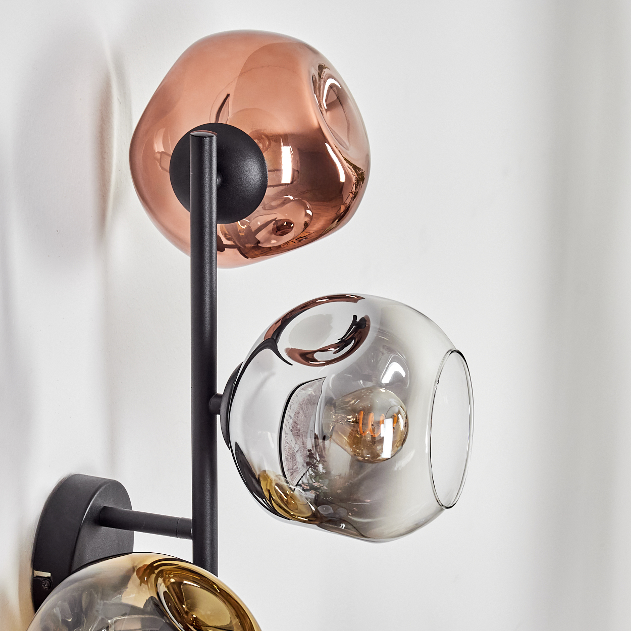 Moderne Wandlampe mit drei Glaskugeln in Kupfer, Grau und Gold. Schwarzer Metallrahmen. Weißer Wandhintergrund.