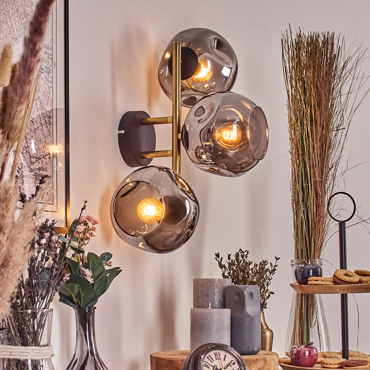 Eine moderne Wandlampe mit drei rauchfarbenen Glaskugeln. Verschiedene Dekorationsartikel, darunter Kerzen und Pflanzen, sind im Vordergrund.