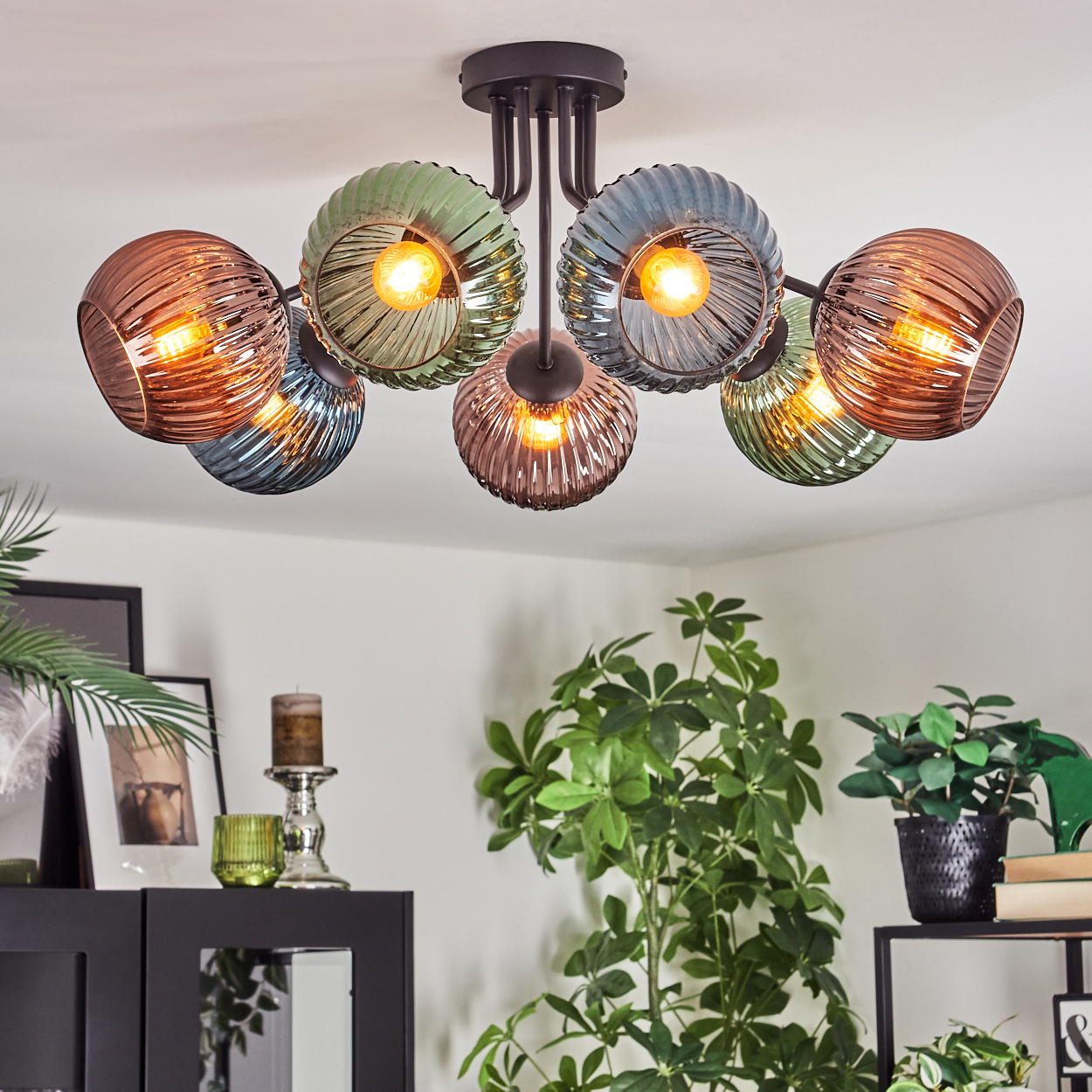Moderne Deckenleuchte mit farbigen Glasschirmen. Pflanzen und schwarze Möbel im Hintergrund. Weiße Wände, helle Beleuchtung.
