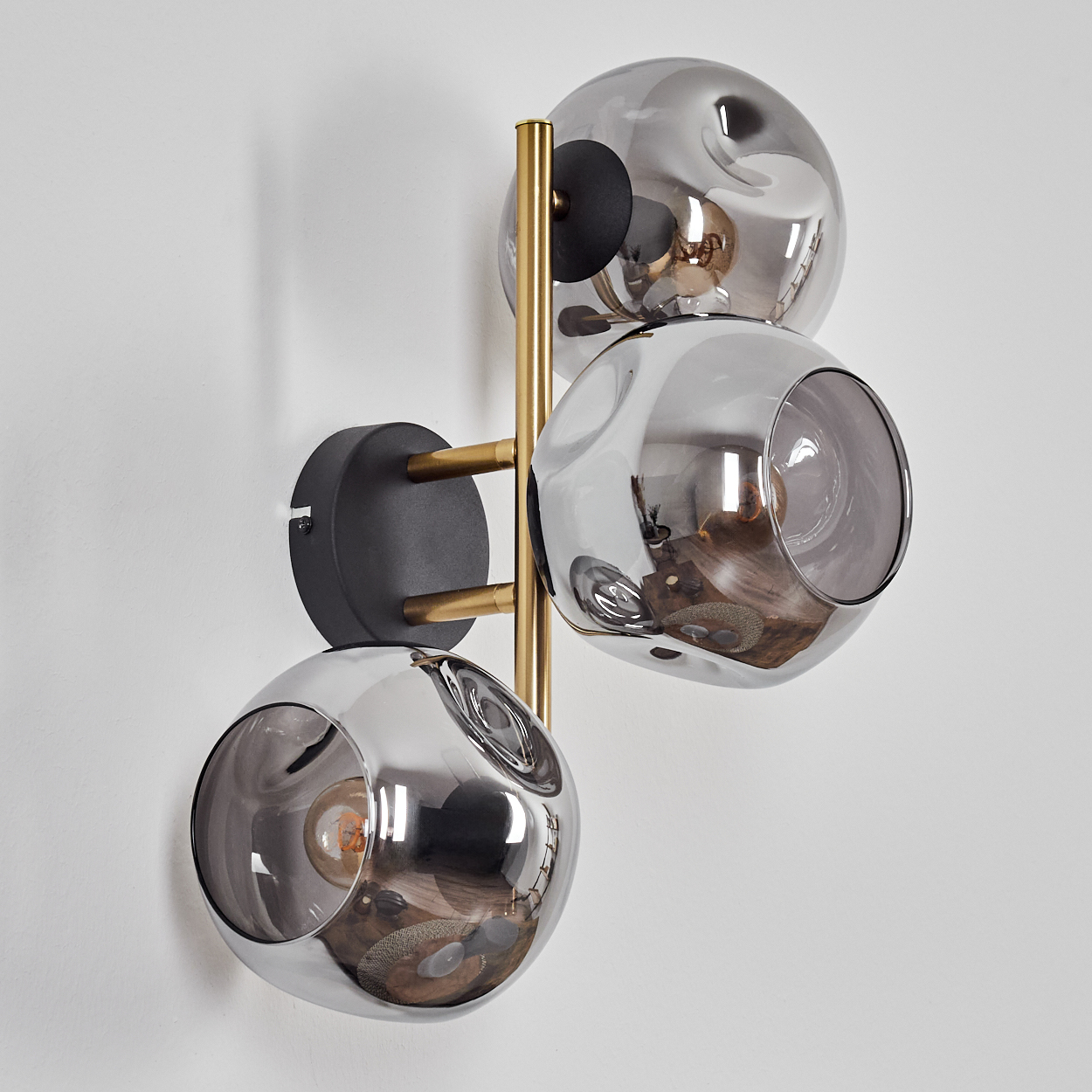 Eine moderne Wandlampe mit drei kugelförmigen, rauchigen Glasschirmen. Sie hat Messingarme und eine schwarze Basis, montiert an einer weißen Wand.