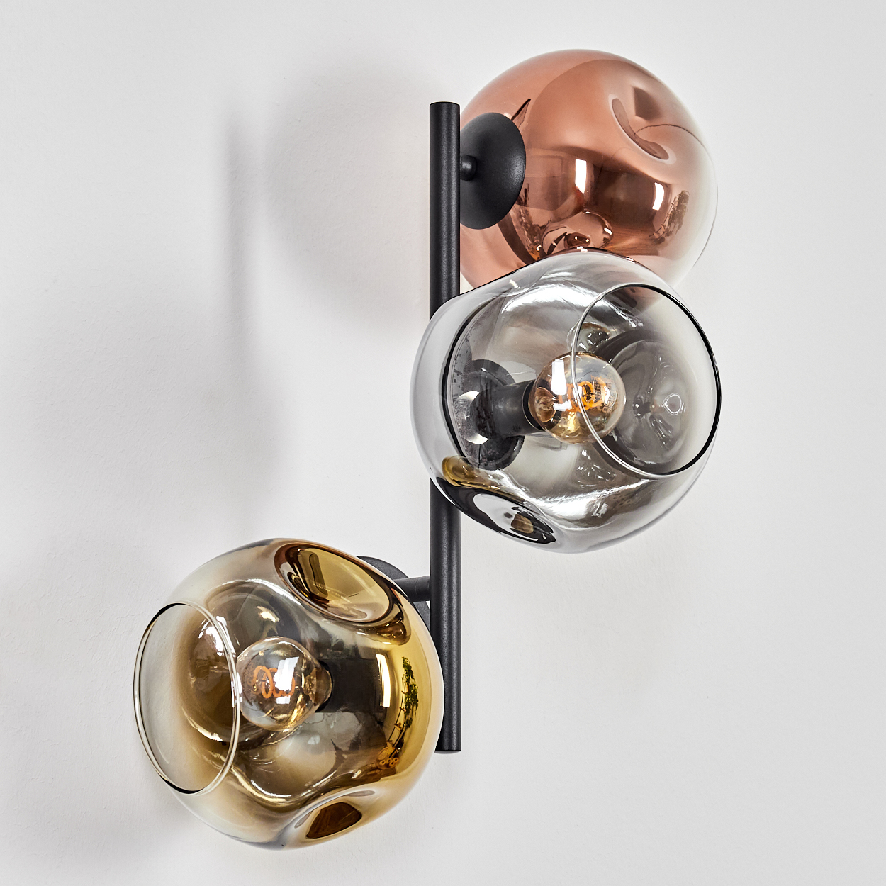 Eine Wandlampe mit drei Glaskugeln in Kupfer-, Grau- und Goldtönen. Jede Kugel hat eine Glühbirne. Schwarze Metallstruktur.
