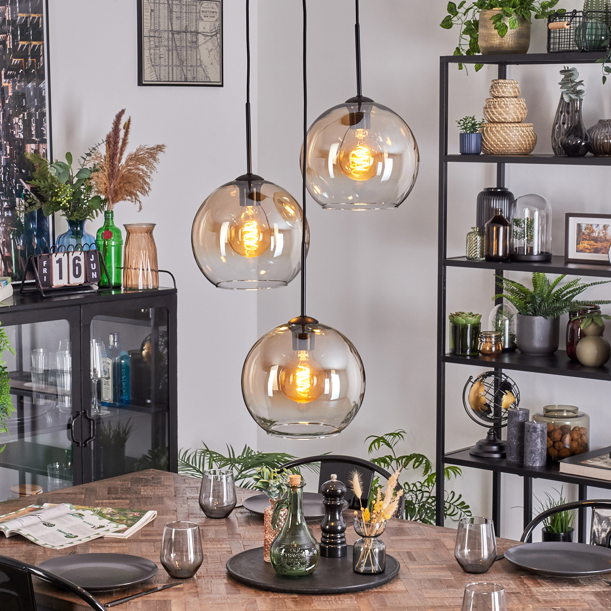 Esszimmer mit drei Hängelampen. Tisch mit Tellern und Gläsern gedeckt. Regale mit Dekoration und Pflanzen im Hintergrund.