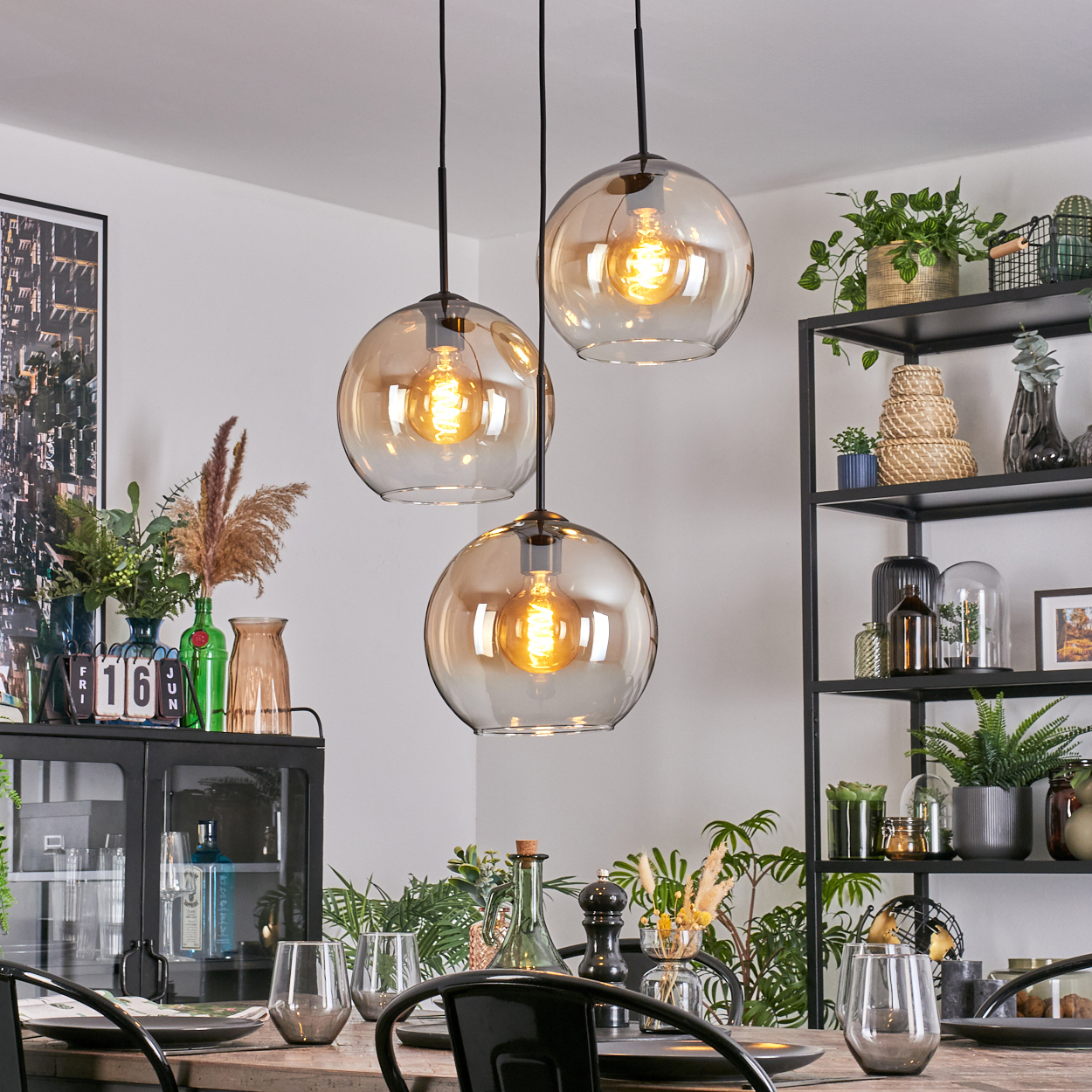 Esszimmer mit drei Hängelampen. Tisch mit Gläsern gedeckt. Regale mit Pflanzen und Dekorationsartikeln.